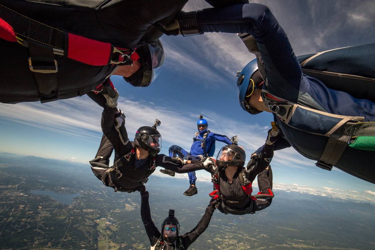 Four experienced skydivers in formation during free fall with one solo skydiver in blue in the background.