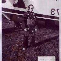 Gentlemen wearing glasses and skydiving gear smiling in front of airplane.