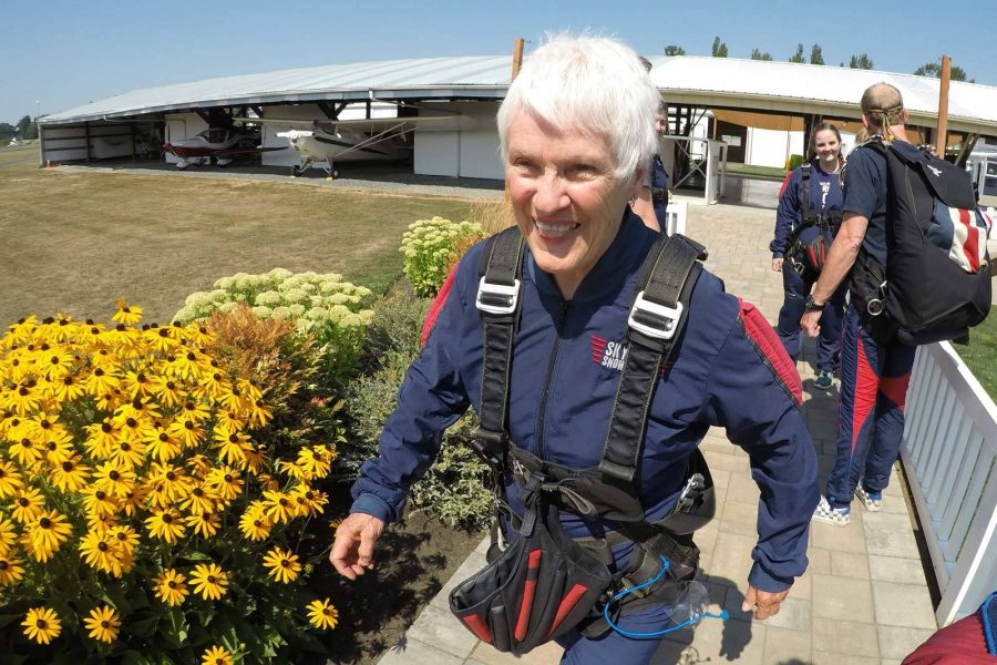 Older female walking past yellow flowers wearing skydiving gear.