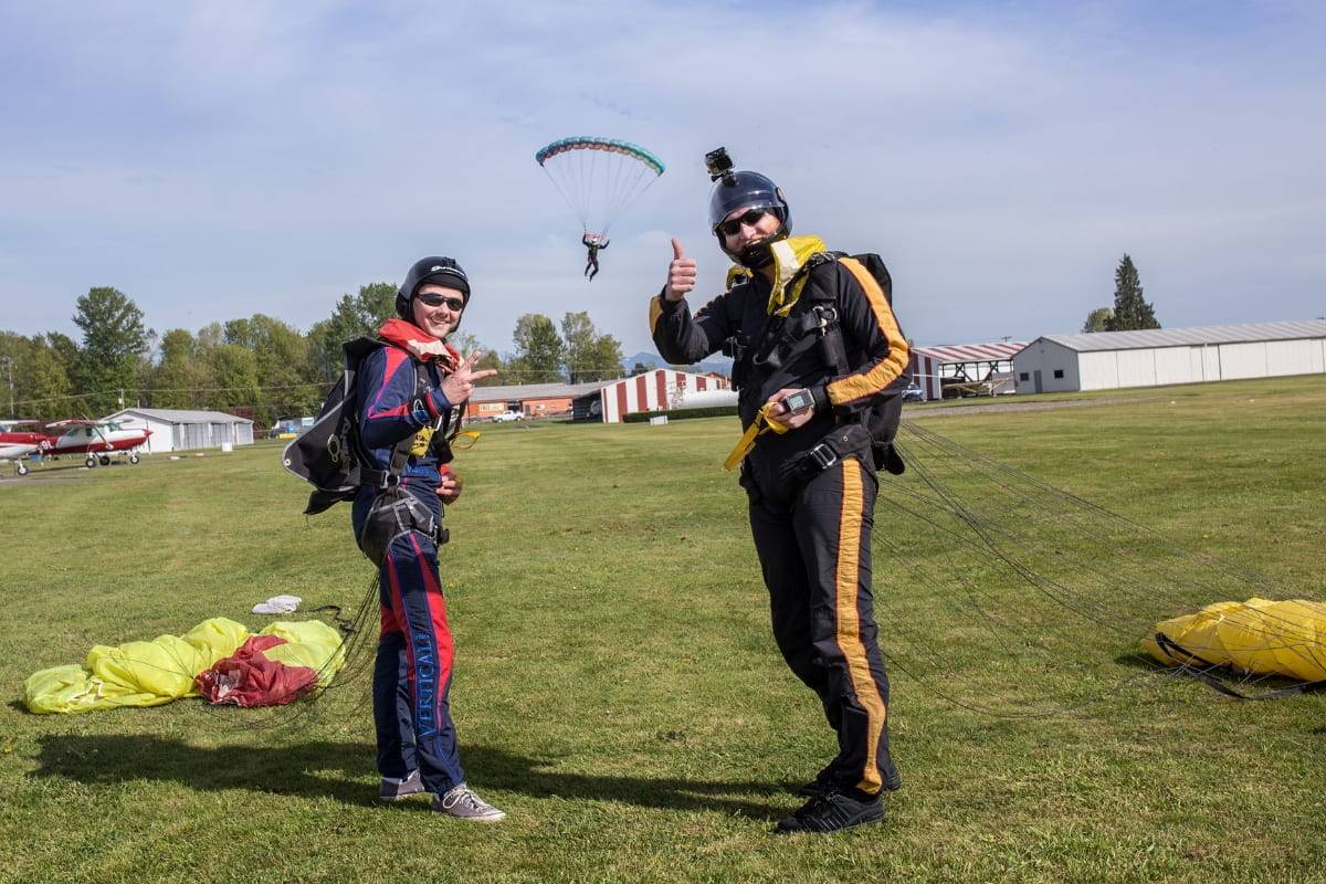 Two skydivers giving thumbs up post skydive with skydiver coming down to land in background.