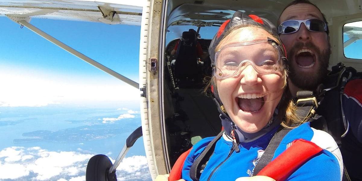 Tandem student wearing blue and red skydiving gear with instructor excited before taking the leap out of the plane.