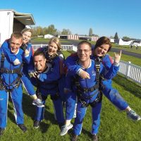 Group of six tandem skydivers wearing all blue gear happily posing outside in grass.