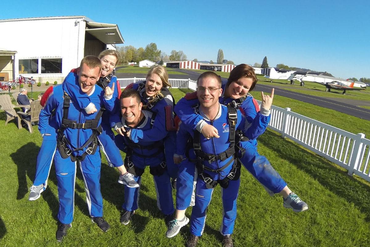 Group of six tandem skydivers wearing all blue gear happily posing outside in grass.