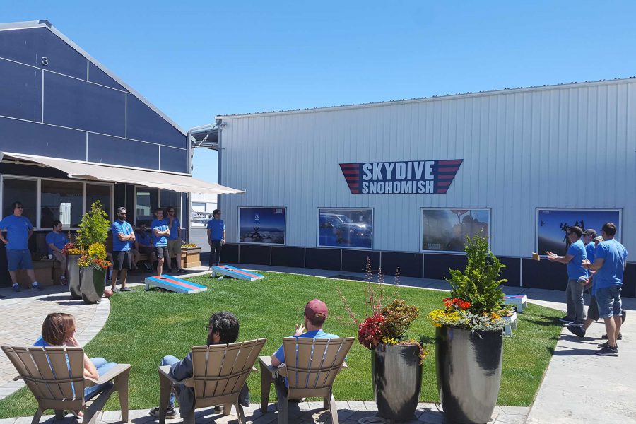 Outside area of Skydive Snohmish with people standing around playing corn hole and sitting in tan chairs.