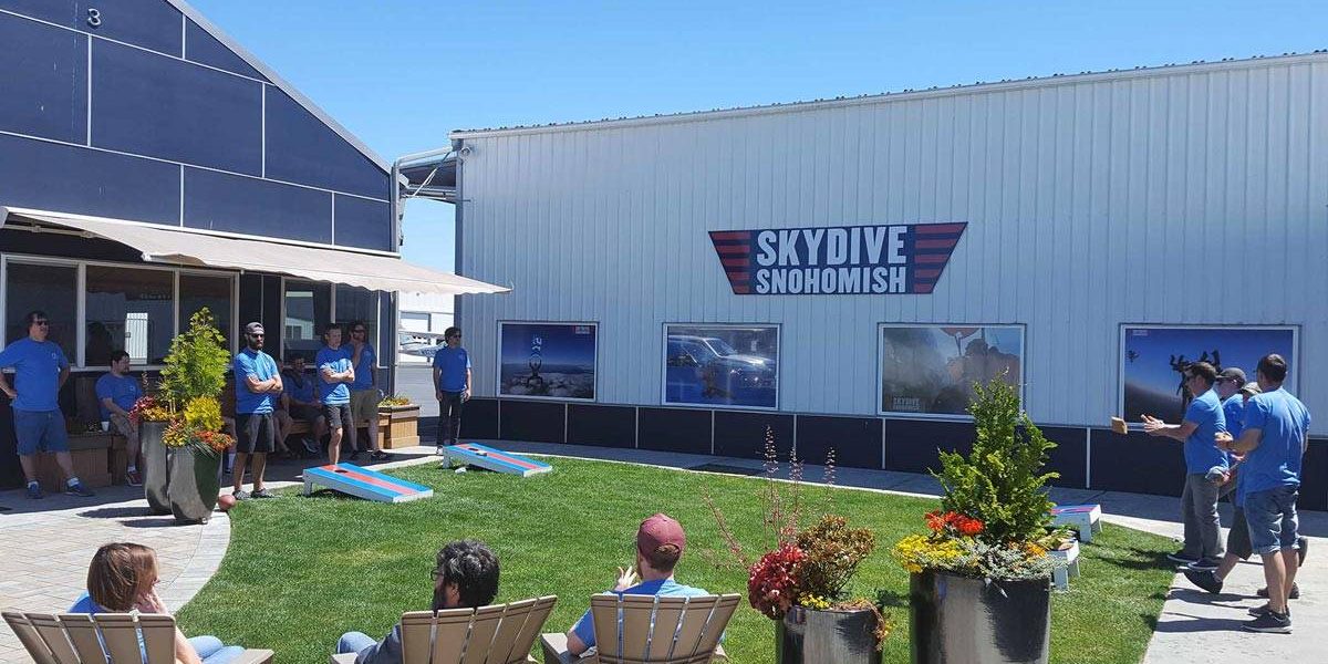 Outside area of Skydive Snohmish with people standing around playing corn hole and sitting in tan chairs.