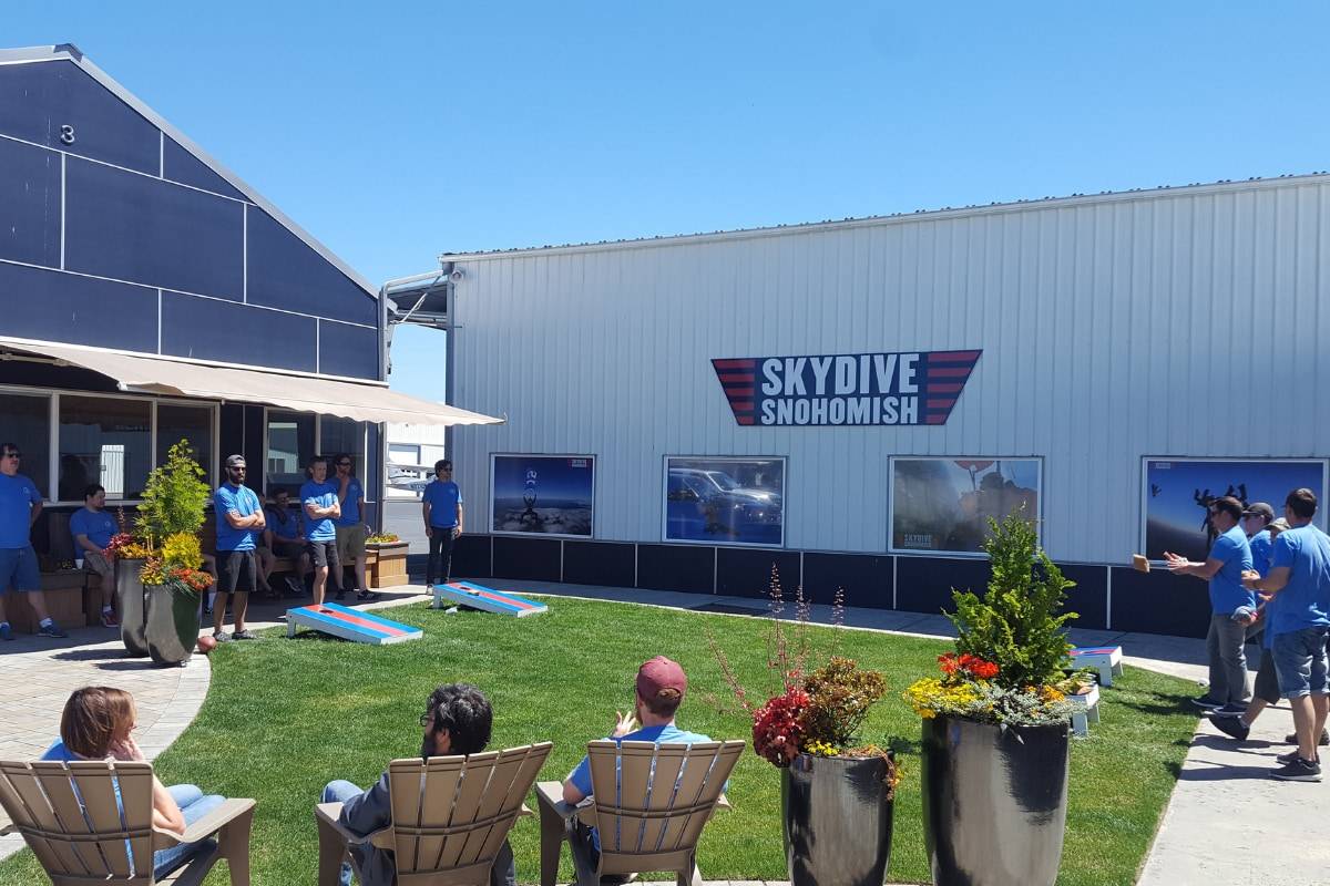 Outside area of Skydive Snohmish with people standing around playing corn hole and sitting in tan chairs.