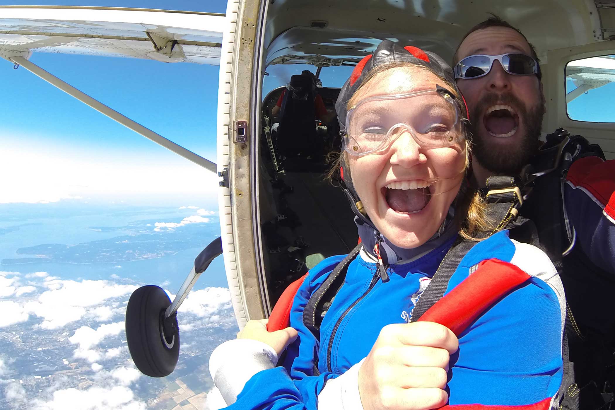 Tandem student wearing blue and red skydiving gear with instructor excited before taking the leap out of the plane.