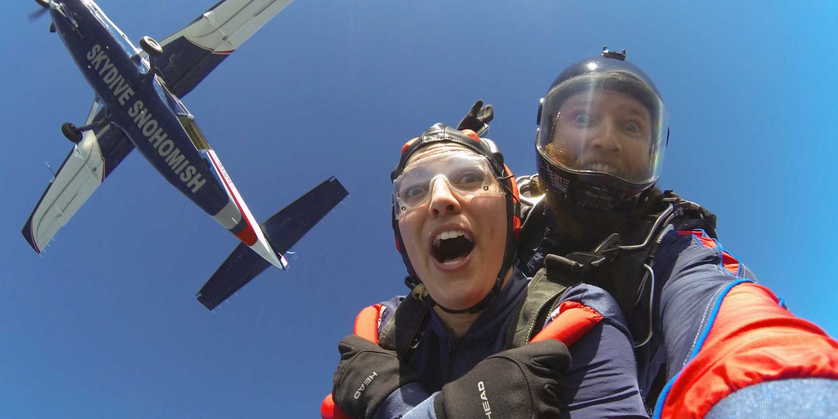 Tandem instructor and female skydiver making excited faces during free fall with blue under belly of plane above them.