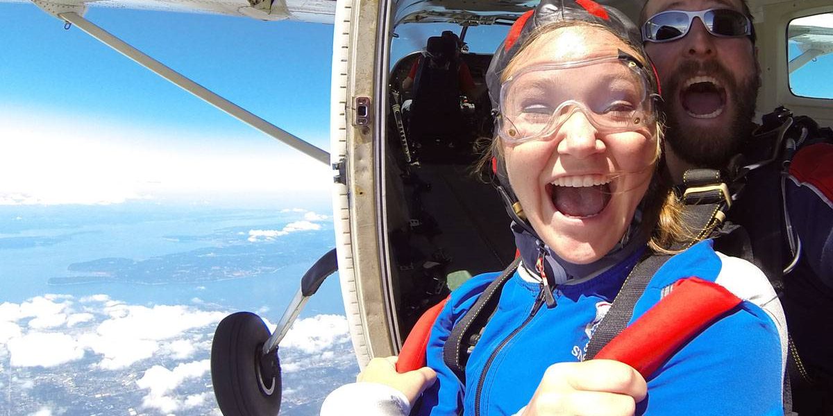 Tandem student wearing blue and red skydiving gear with instructor excited before taking the leap out of the plane.