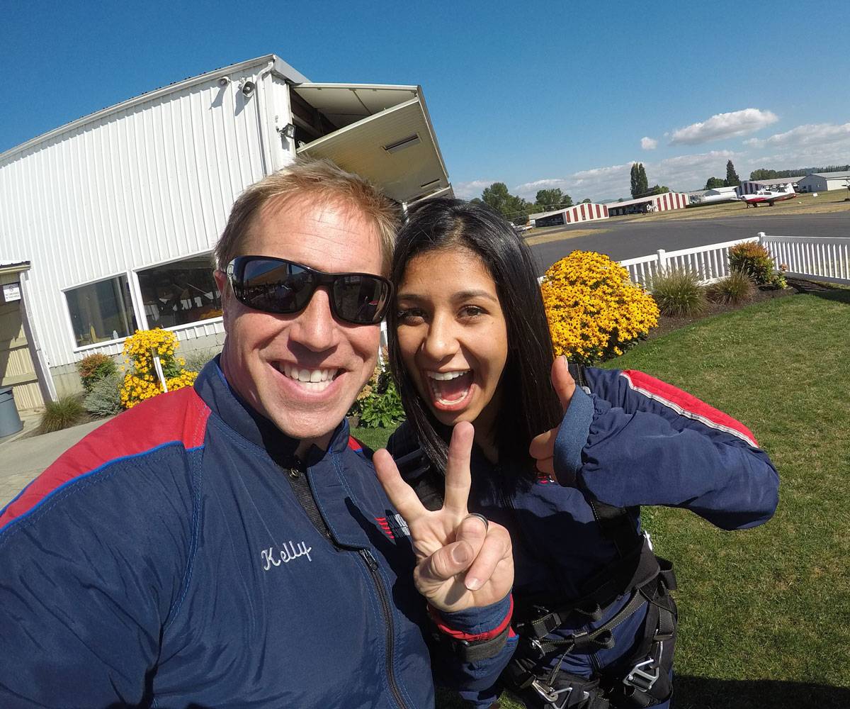 Female tandem skydiver with black hair smiling and giving thumbs up with tandem instructor.