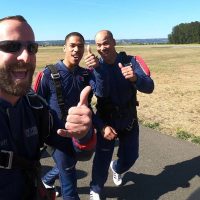 Three male skydivers walking towards airplane to skydive giving thumbs up.