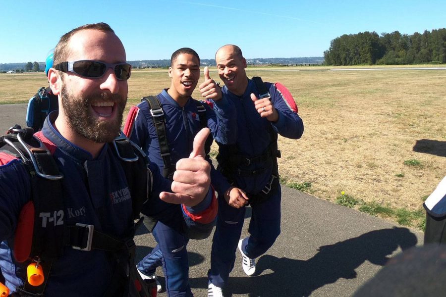 Three male skydivers walking towards airplane to skydive giving thumbs up.
