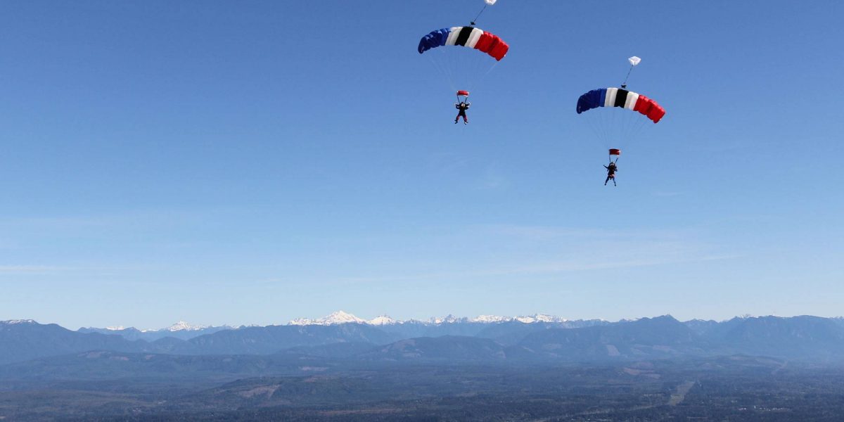 Two experienced skydivers coming towards land with mountains in the back ground and white, blue, red, and black canopies.