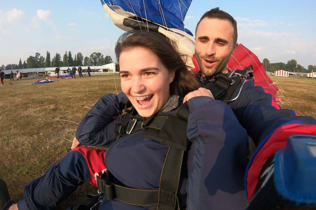 Young women in braces smiling post landing while wearing blue skydiving gear.